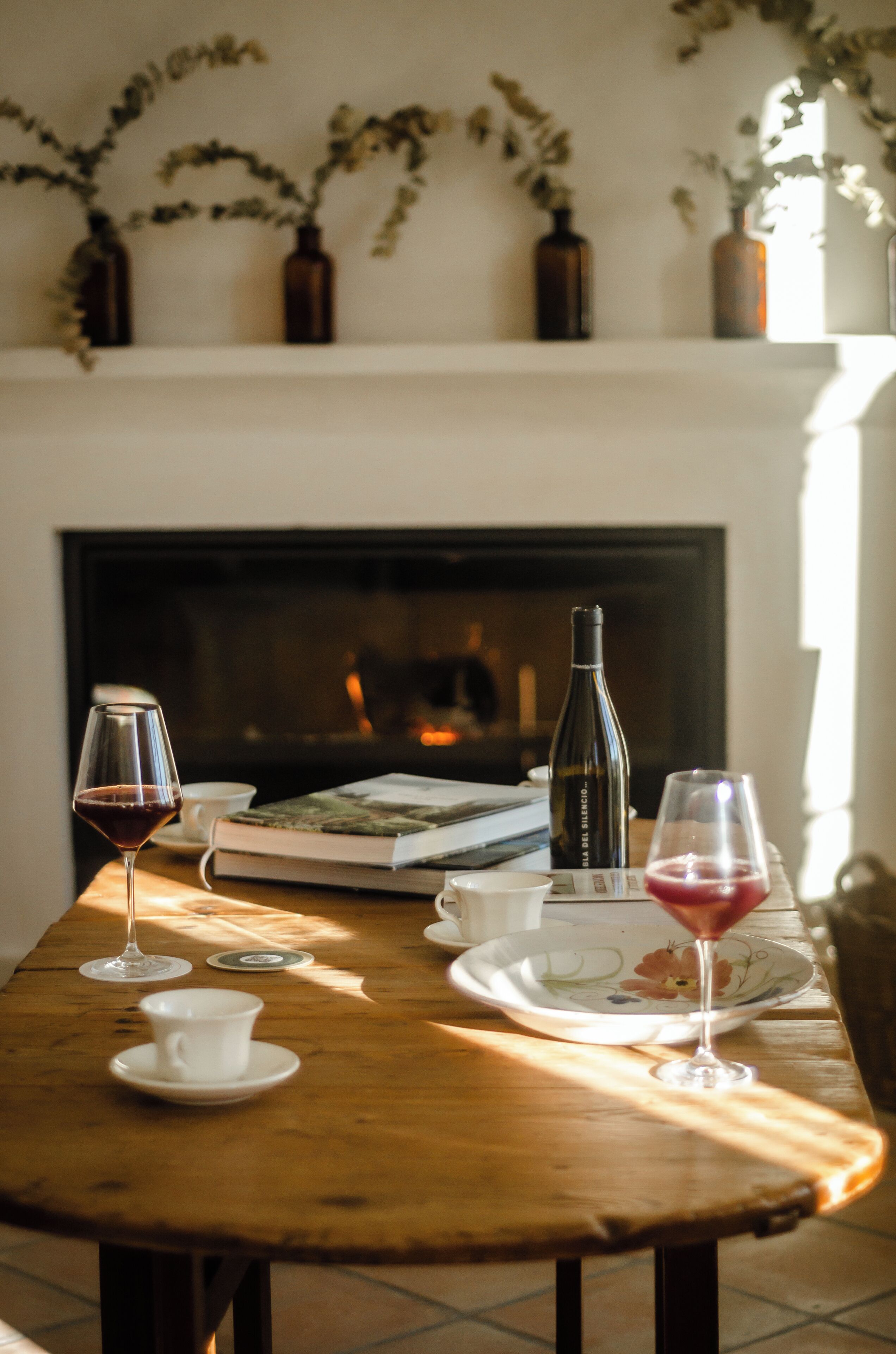 A wooden table with wine glasses, a bottle, and a book in a cozy room with a fireplace.