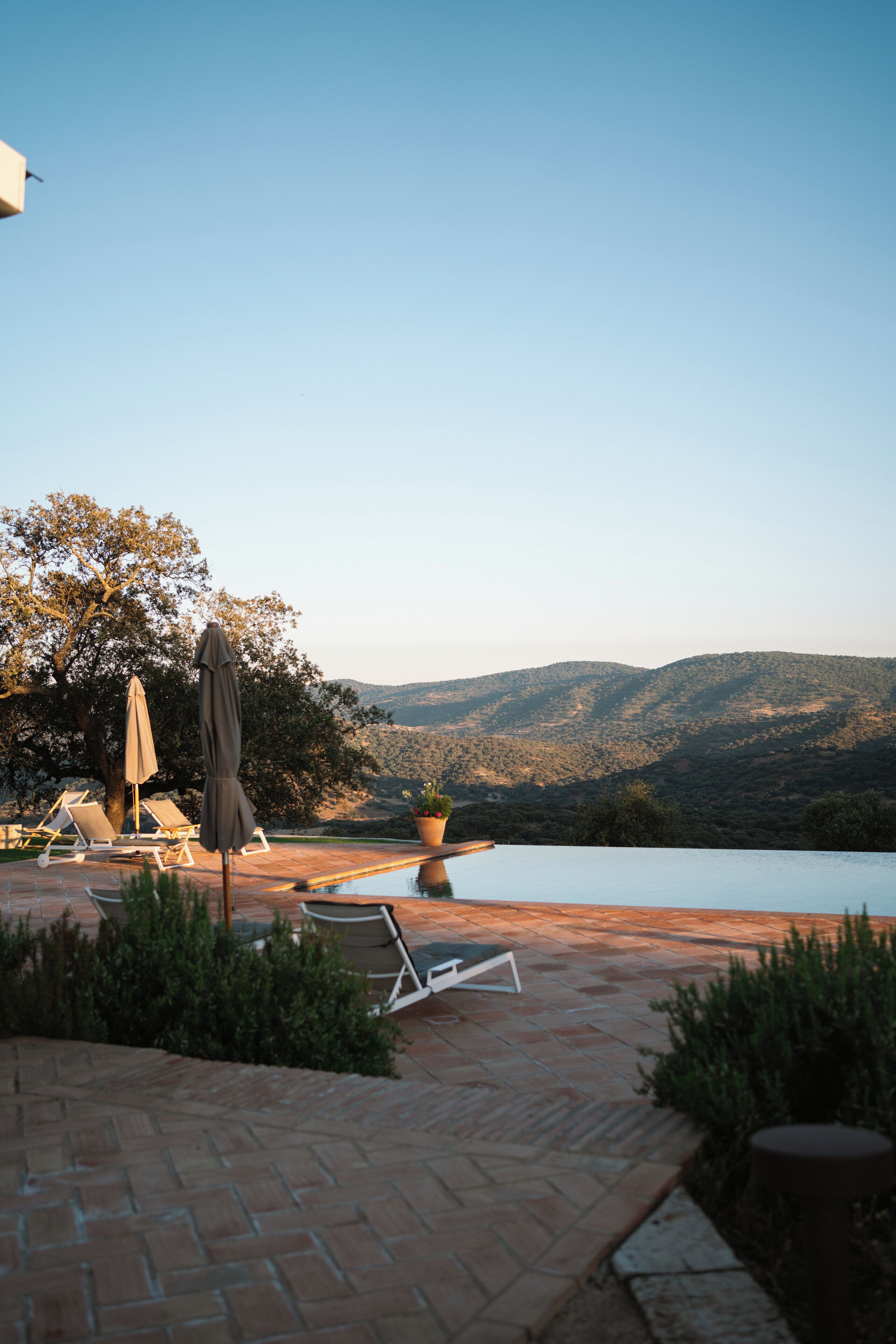 Un espace aménagé au bord de la piscine, avec des chaises longues et un parasol, entouré d’une terrasse en briques et offrant une vue sur les collines.