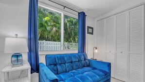 Une pièce avec un canapé bleu, une table d’appoint blanche avec une lampe, une fenêtre avec des rideaux bleus et une armoire blanche.