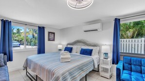 Une chambre d’hôtel moderne avec un grand lit, une lampe sur pied, un fauteuil bleu et une vue sur des palmiers.