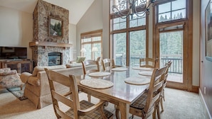 A spacious living room with a fireplace, a dining area with a large table and chairs, and large windows offering a view of the outdoors.