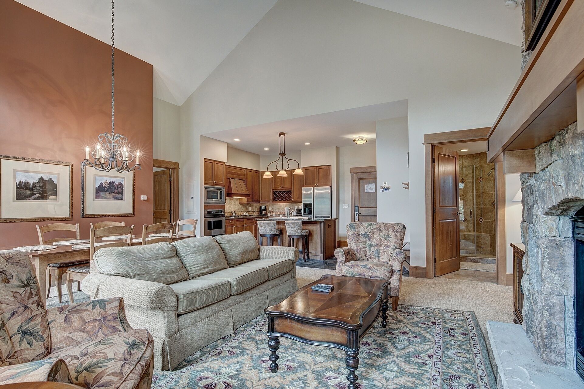 A spacious living room with a sofa, armchairs, and a coffee table. There is a kitchen area with wooden cabinets and a dining table.