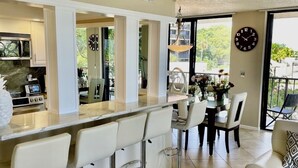 Une cuisine moderne dotée d’un îlot central, de tabourets de bar, d’un coin repas avec une table et des chaises, et d’une grande fenêtre offrant une vue sur la verdure.