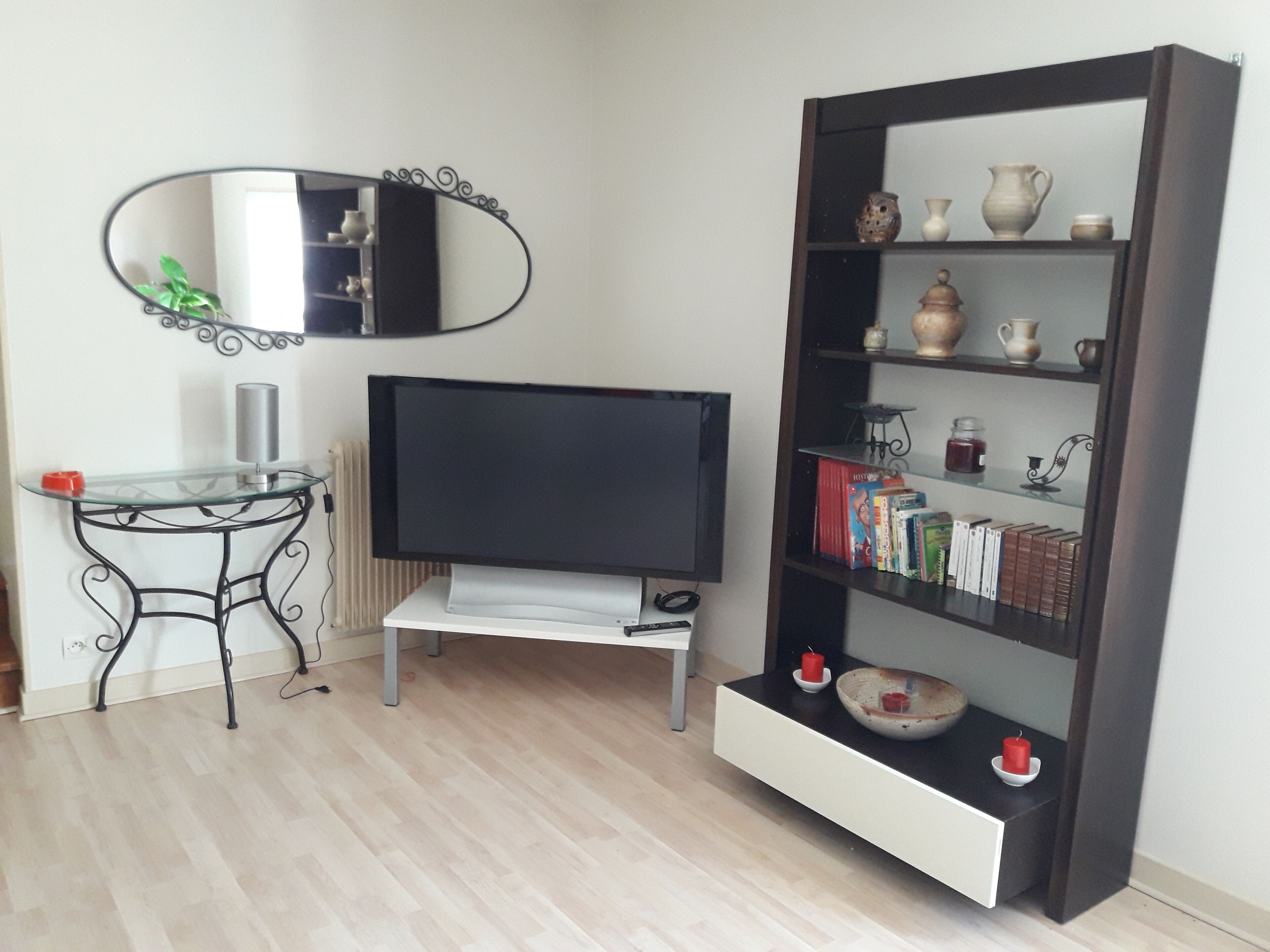 A living room with a flat-screen TV on a stand, a glass-top coffee table, a black bookshelf with decorative items, and a wall-mounted mirror.