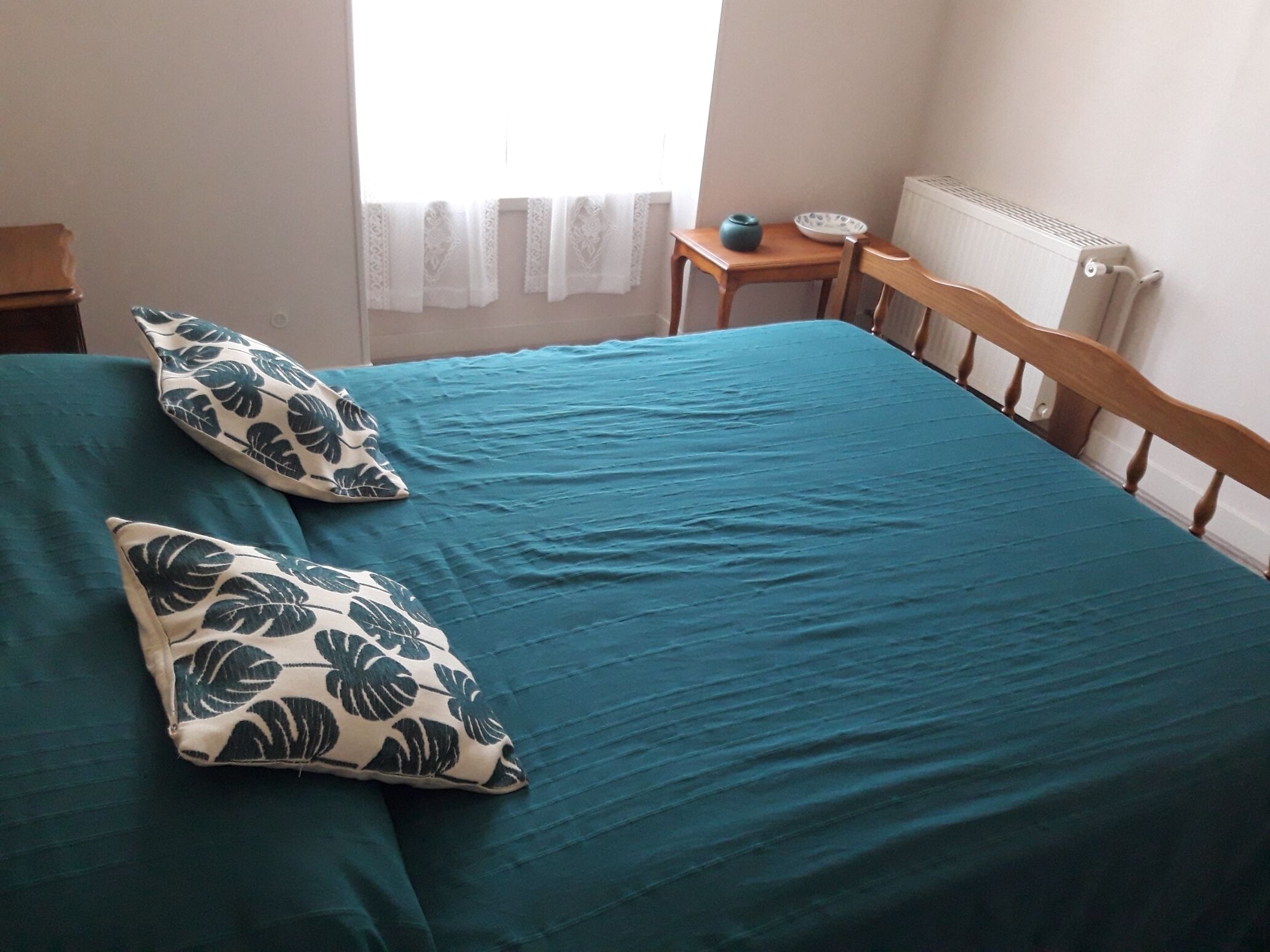 A bedroom with a bed, two pillows, a nightstand, a small table, and a radiator.