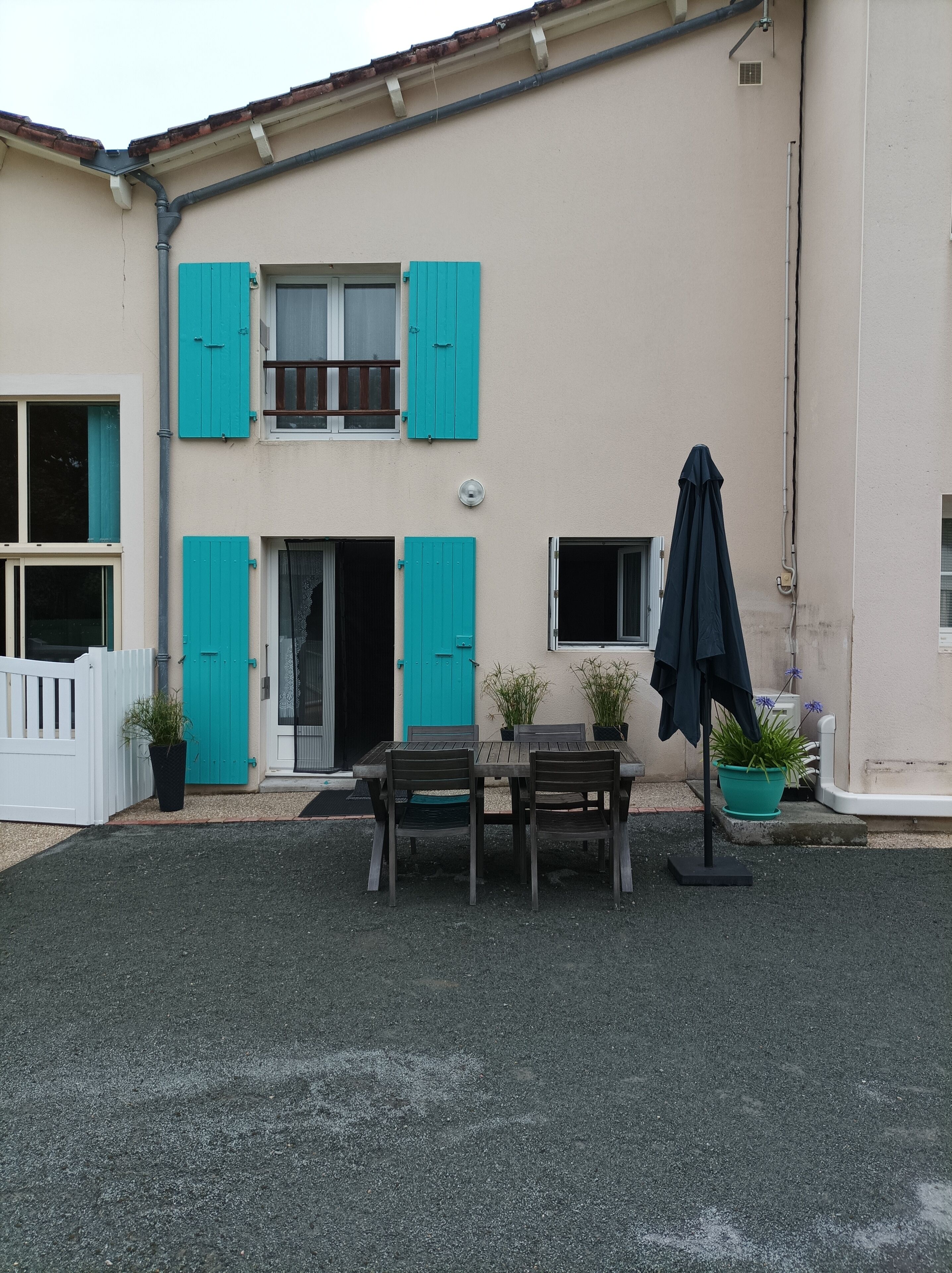 Een gebouw met twee verdiepingen, voorzien van blauwgroene luiken, een balkon en een klein terras met een tafel en stoelen.