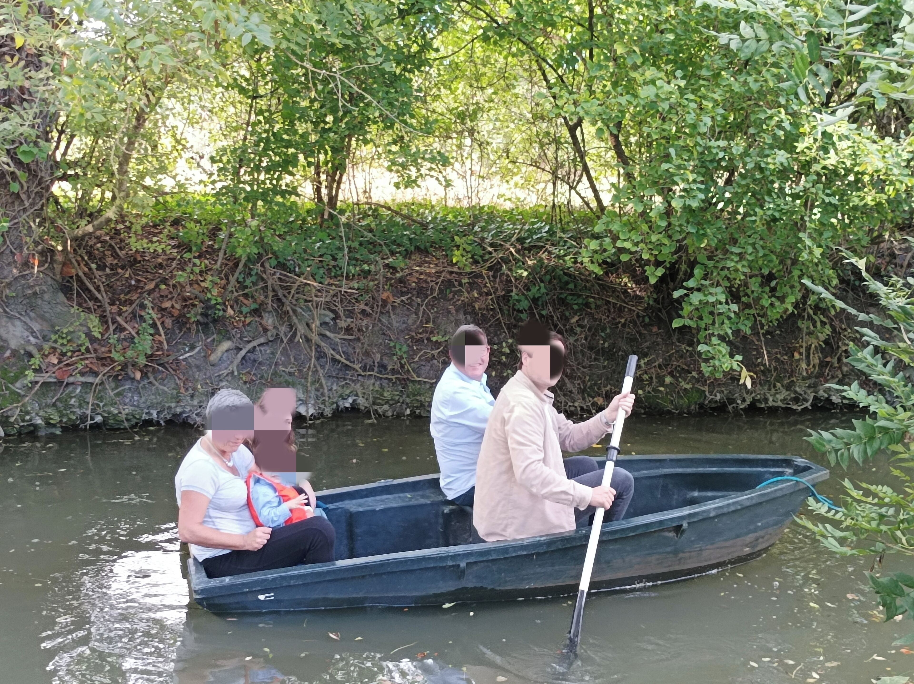 A narrow boat with three passengers, one holding a paddle, on a calm river surrounded by dense greenery.