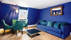 Una stanza con una parete blu, un divano blu con cuscini decorati, una poltrona verde, un tavolino da caffè nero e uno specchio con cornice dorata.