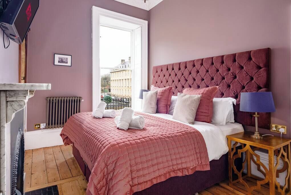 A bedroom with a large bed, a wooden nightstand, a marble fireplace, and a view of buildings outside.