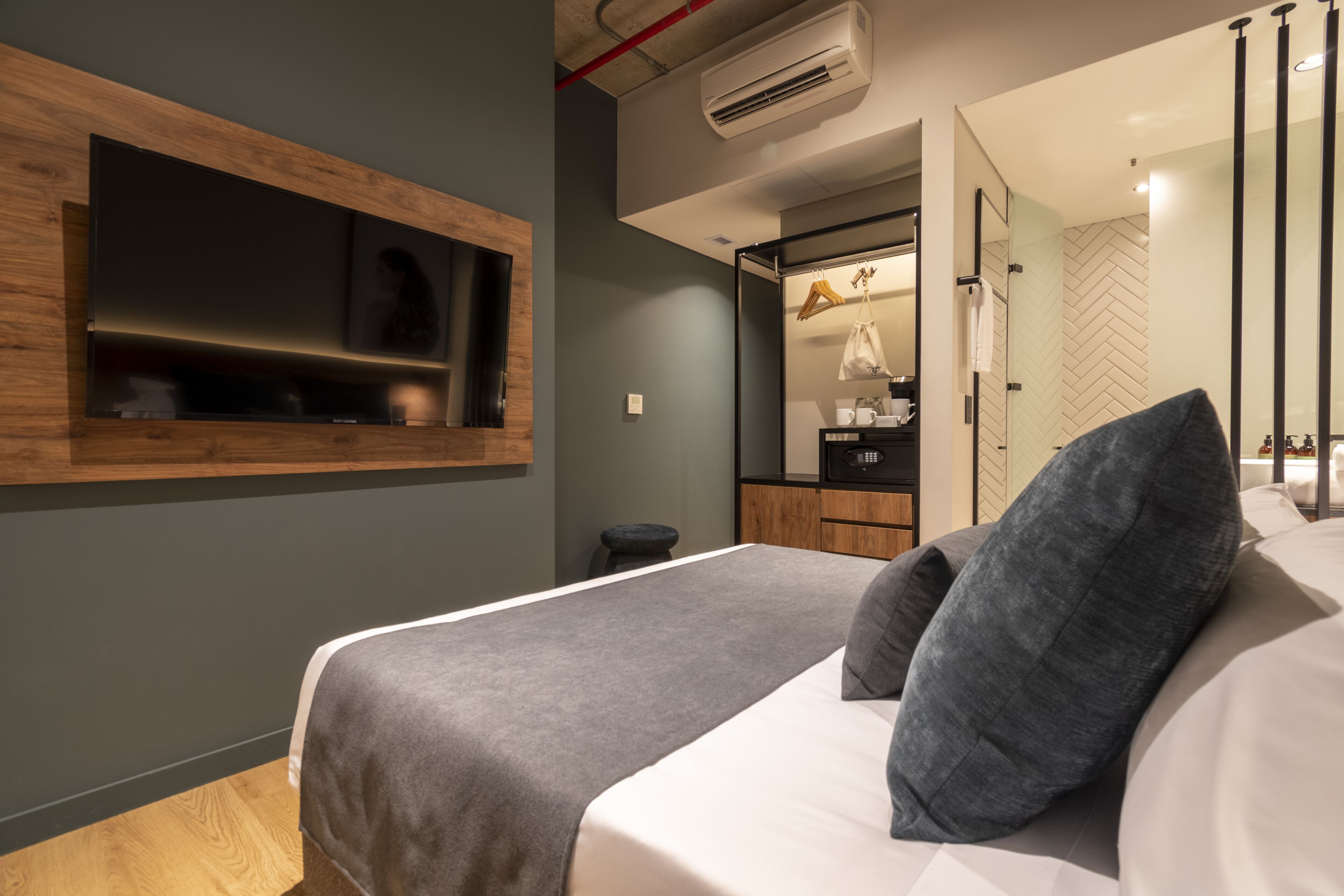 Moderni hotellihuone, jossa on suuri sänky, seinään upotettu televisio, pieni jakkara ja peili, jonka takana on kalanruotokuviolla koristeltu seinä.