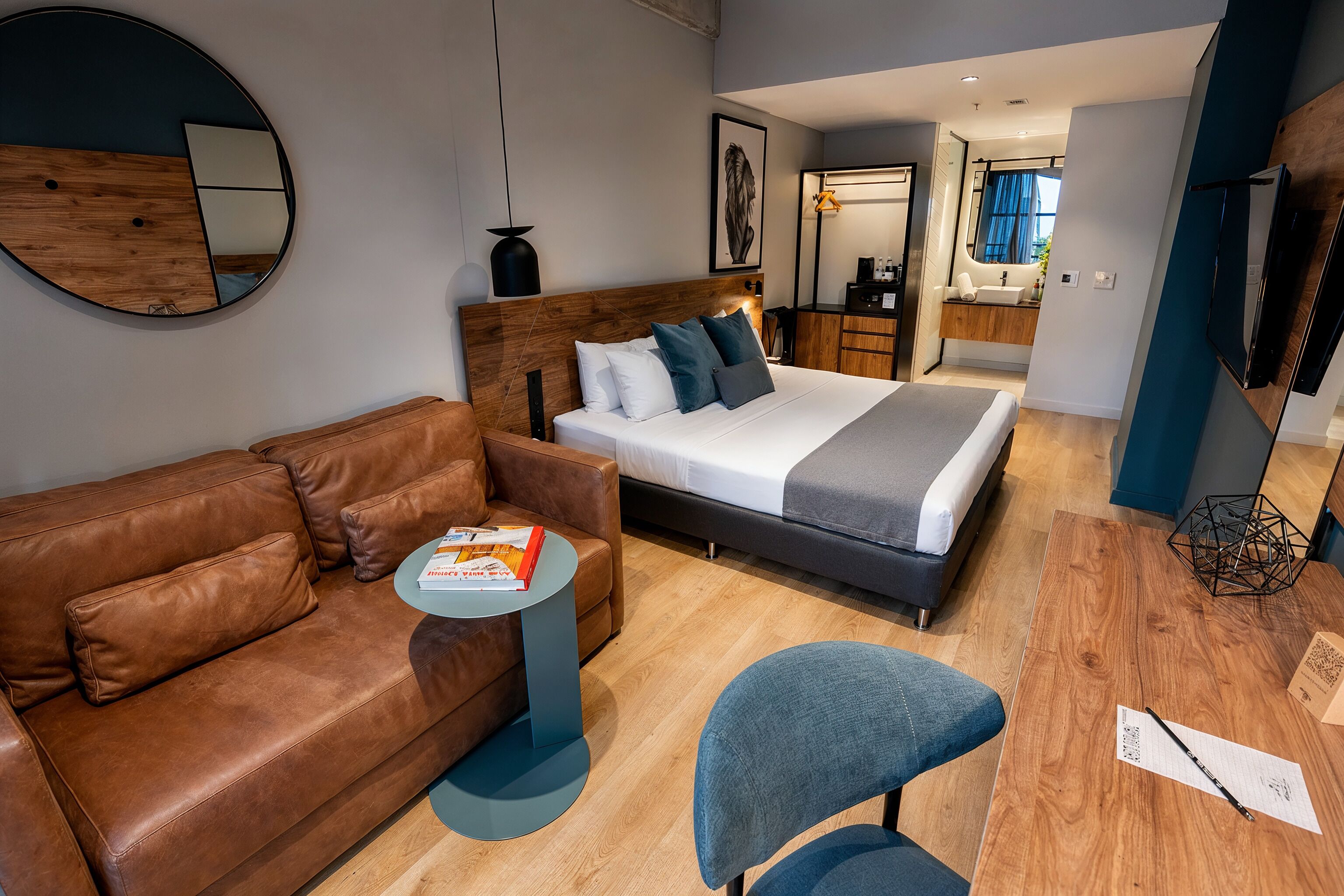 Een moderne hotelkamer met een bed, een bruine leren bank, een klein rond tafeltje, een blauwe stoel en een houten vloer.