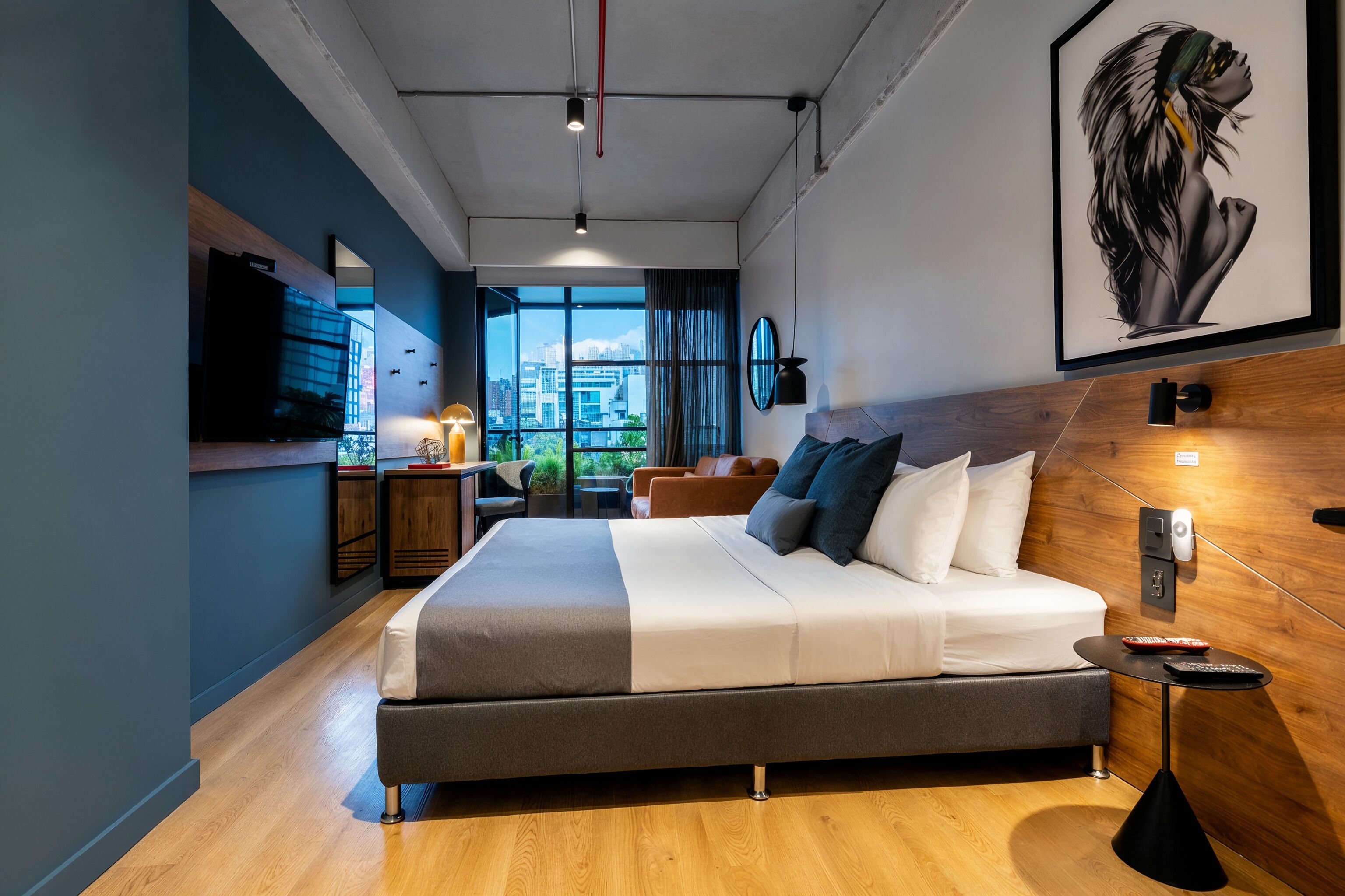 A modern hotel room with a large bed, a desk, a chair, a TV, and a city view.