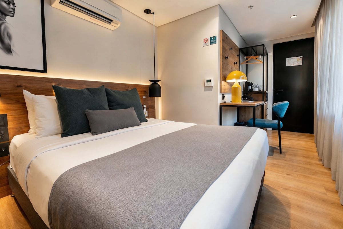 A modern hotel room with a large bed, bedside table, lamp, and a desk with a chair.