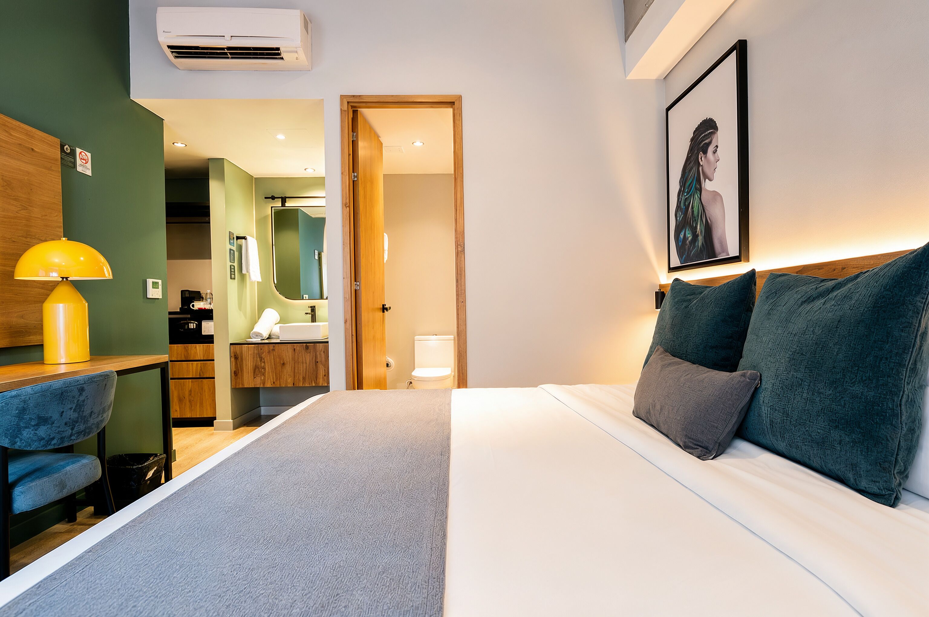 A hotel room with a bed, a desk with a lamp, a bathroom, and a hallway leading to another room.