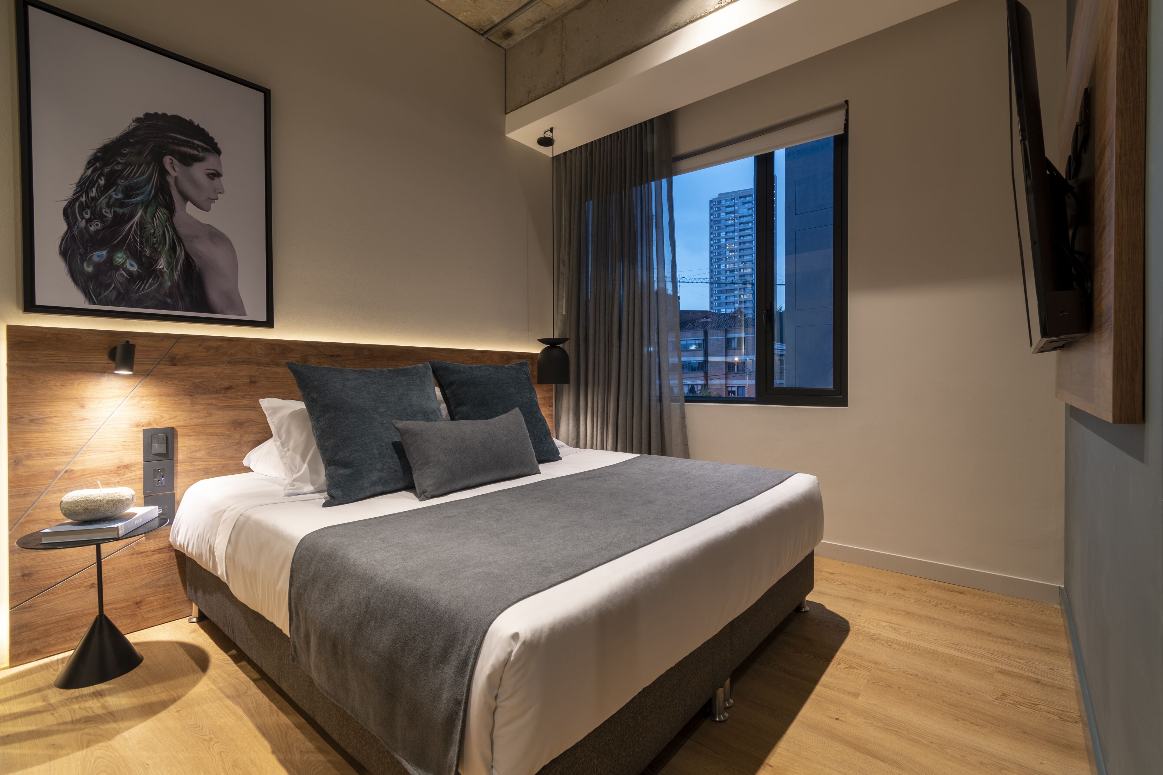 A modern hotel room with a large bed, a bedside table with a lamp, a mounted TV, and a window with a city view.