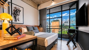 Una habitación de hotel moderna con una cama grande, un escritorio con lámpara, una silla, una estantería y un ventanal con vistas a la ciudad.