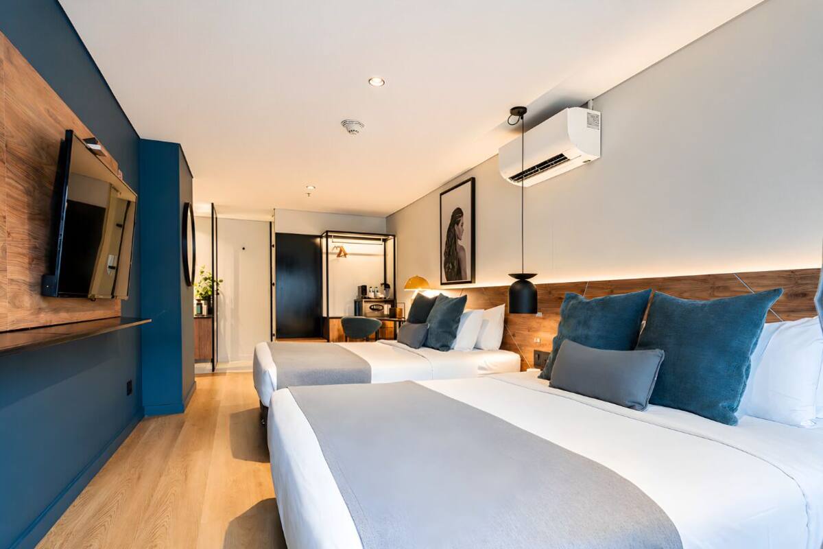 A modern hotel room with two beds, a flat-screen TV, a wooden accent wall, and a ceiling-mounted air conditioner.