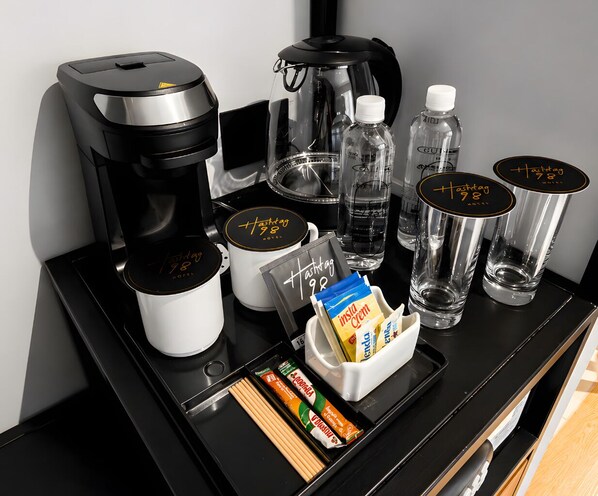 A hotel minibar setup with a coffee maker, a glass carafe, a water bottle, a glass of water, a glass of juice, and a tray with snacks.