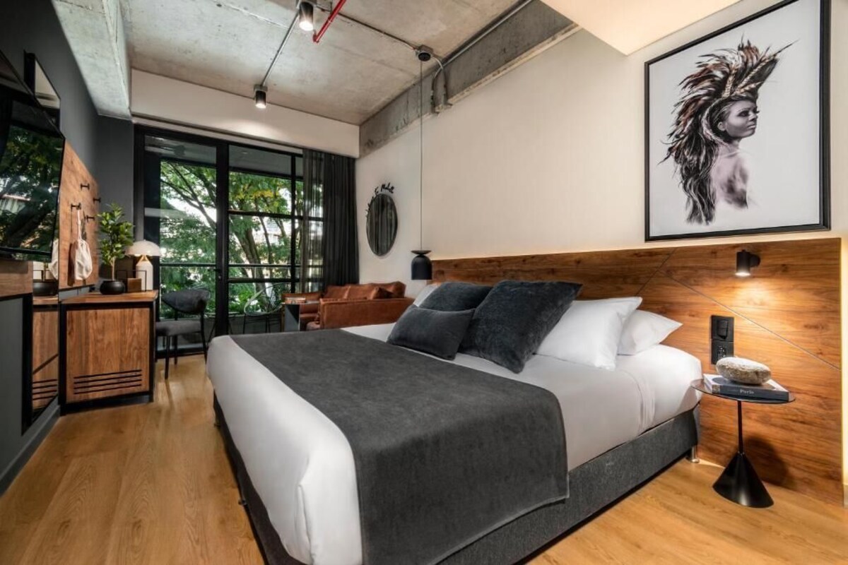 A modern bedroom with a large bed, wooden furniture, a desk, and a view of greenery.