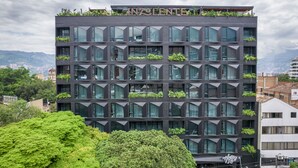 Un edificio moderno con fachada geométrica, varios balcones con vegetación y vistas claras del paisaje urbano circundante.