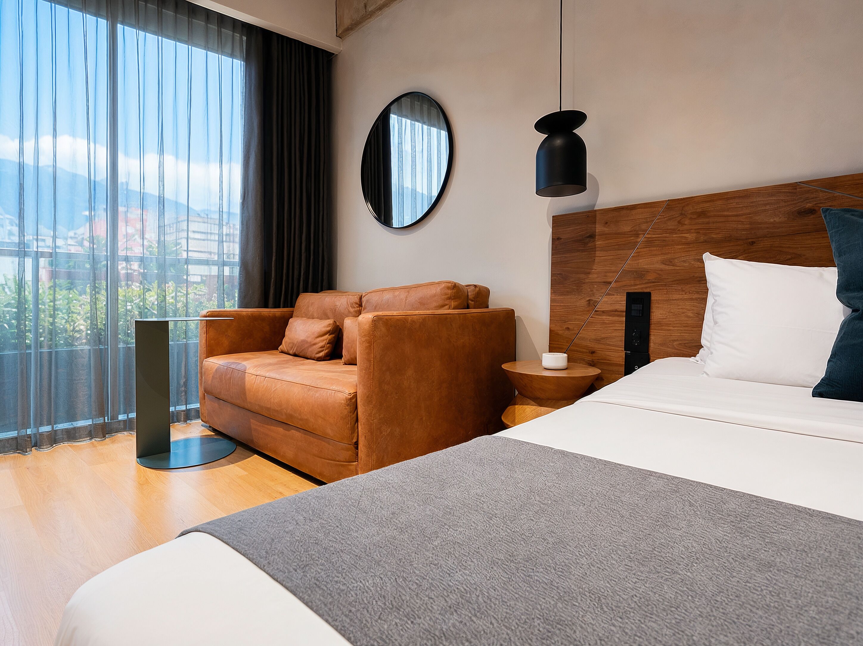 Una habitación de hotel moderna con una cama grande, un sillón de cuero, un espejo redondo, un cabecero de madera y vistas a la ciudad.