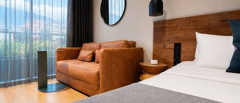Una habitación de hotel moderna con una cama grande, un sillón de cuero, un espejo redondo, un cabecero de madera y vistas a la ciudad.