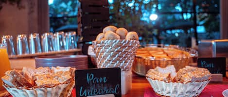 Un bufé de desayuno con varios panes y pasteles expuestos en cestas, un cartel que indica 'Pan Integral' y un letrero que anuncia 'Amena Cocina Consistente'.