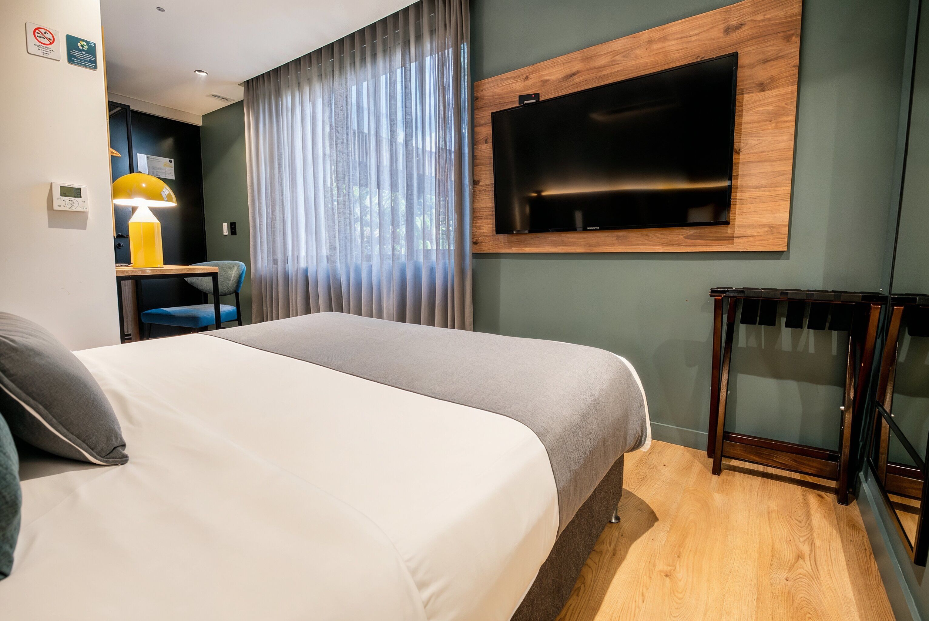 A hotel room with a bed, a desk with a lamp, a TV mounted on the wall, and a window with curtains.