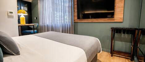 A hotel room with a bed, a desk with a lamp, a TV mounted on the wall, and a window with curtains.