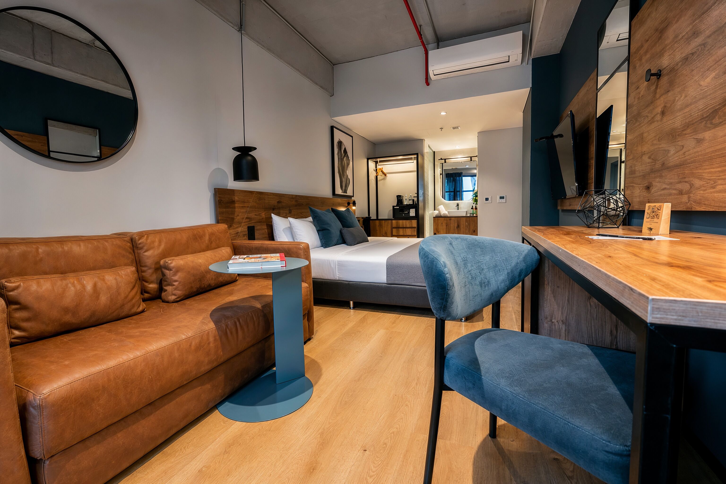 A modern room with a brown leather sofa, a small round table, a blue chair, a bed with a wooden headboard, and a wooden desk with a mirror.