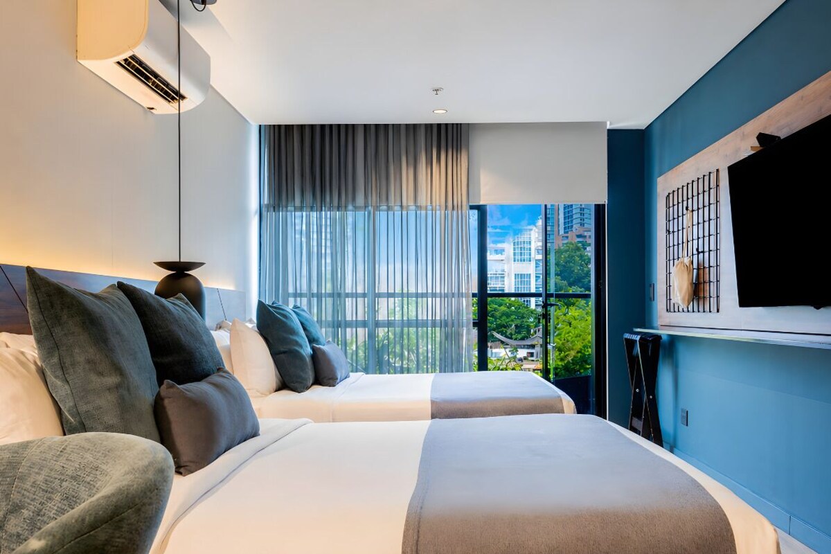 A modern hotel room with two beds, a large window with curtains, a TV mounted on the wall, and an air conditioning unit.