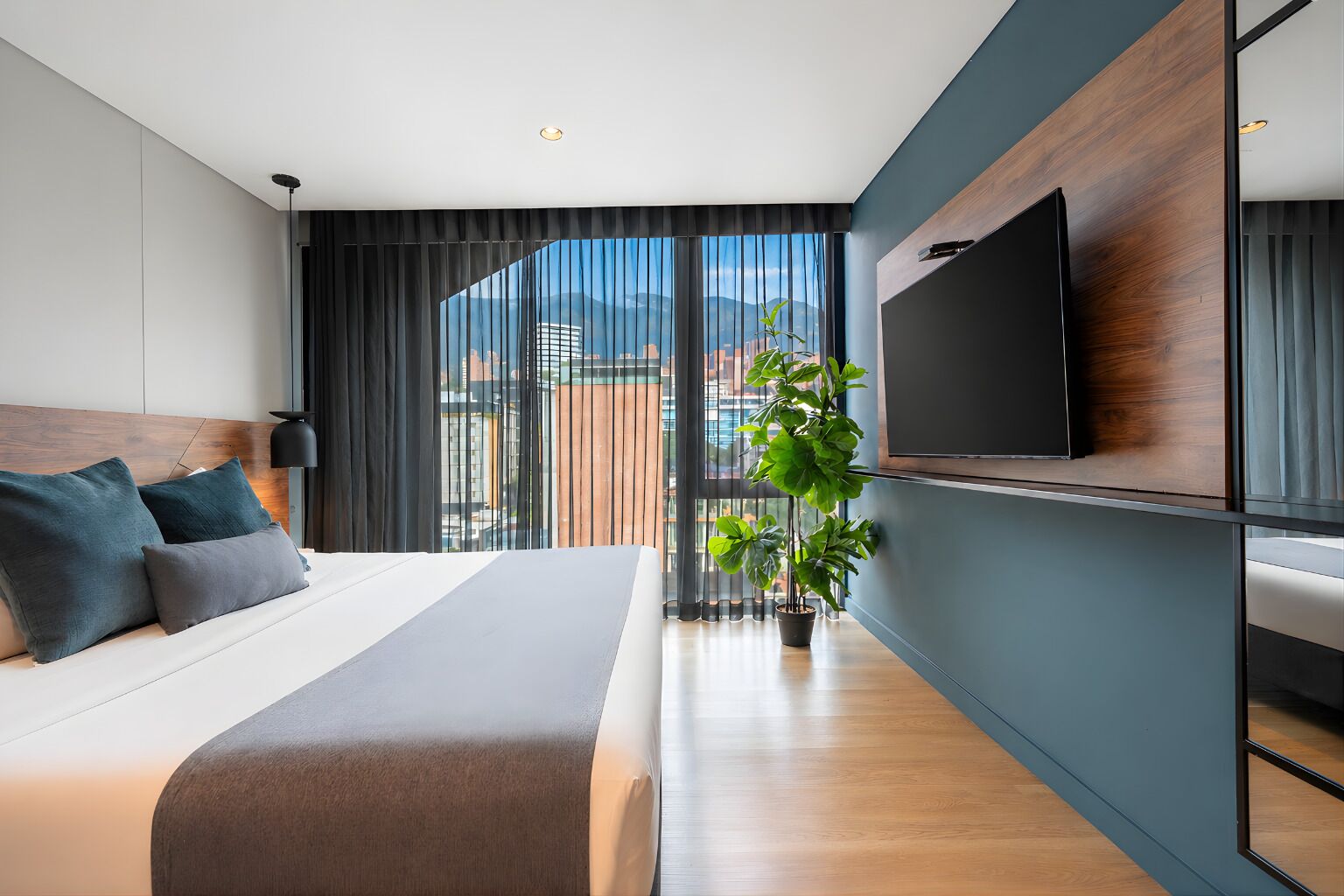 Quarto de hotel moderno com uma cama grande, televisão de tela plana na parede, uma planta em vaso e vista para o horizonte da cidade pela janela.