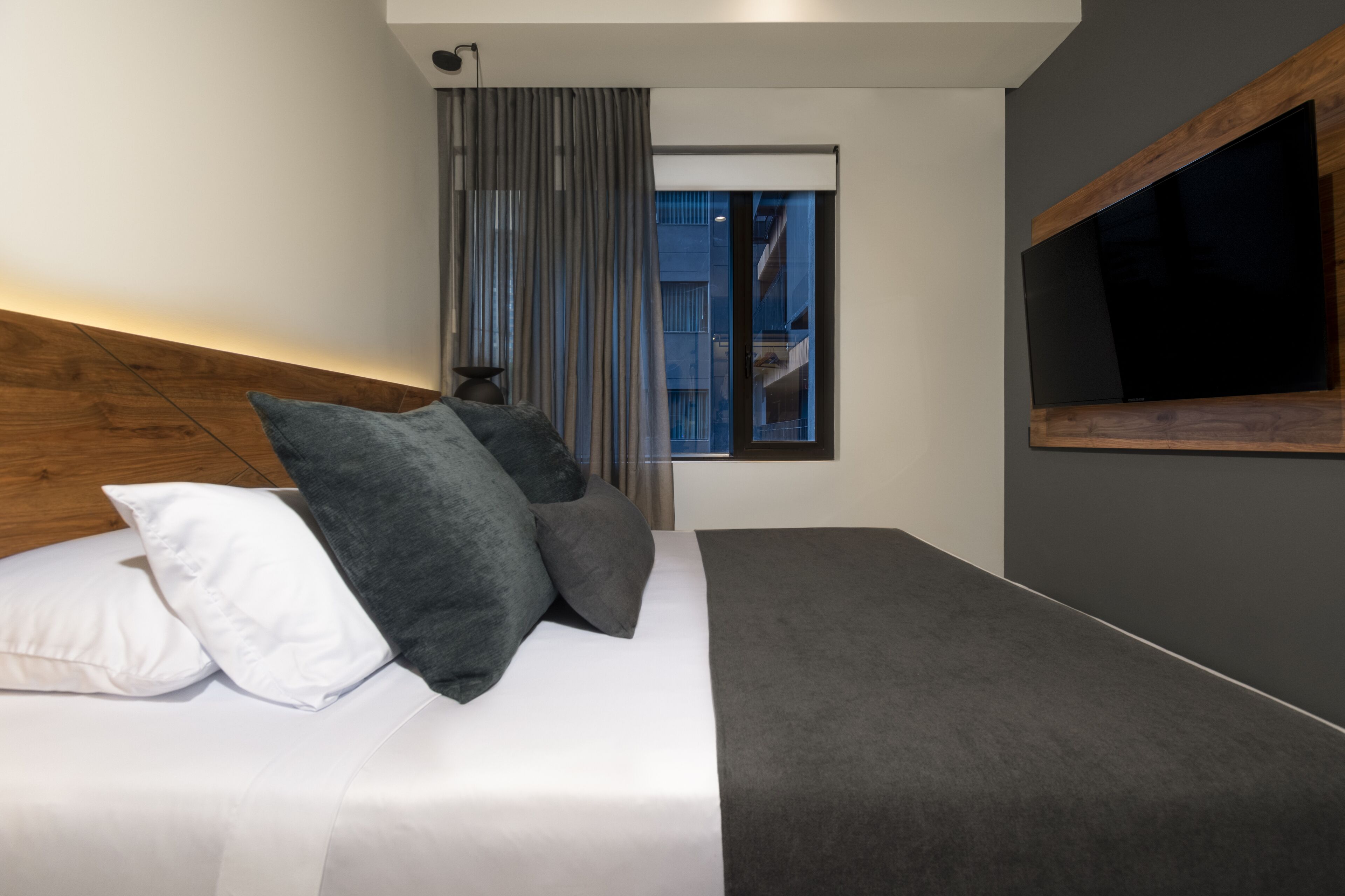 A modern hotel room with a large bed, a flat-screen TV mounted on the wall, and a window with curtains.