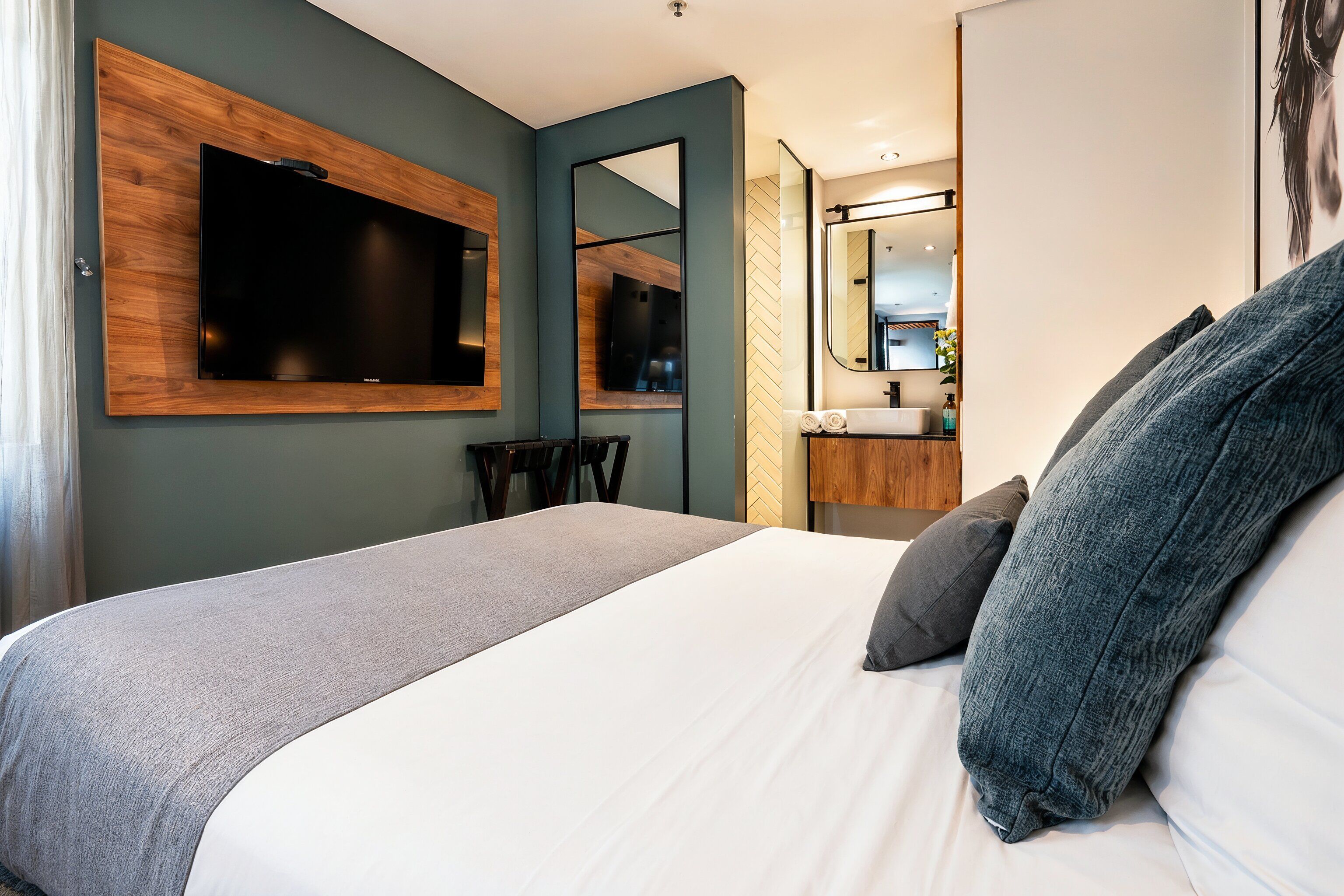 Une chambre d’hôtel moderne avec un grand lit, un téléviseur fixé au mur, un miroir et une salle de bain visible à travers une porte ouverte.