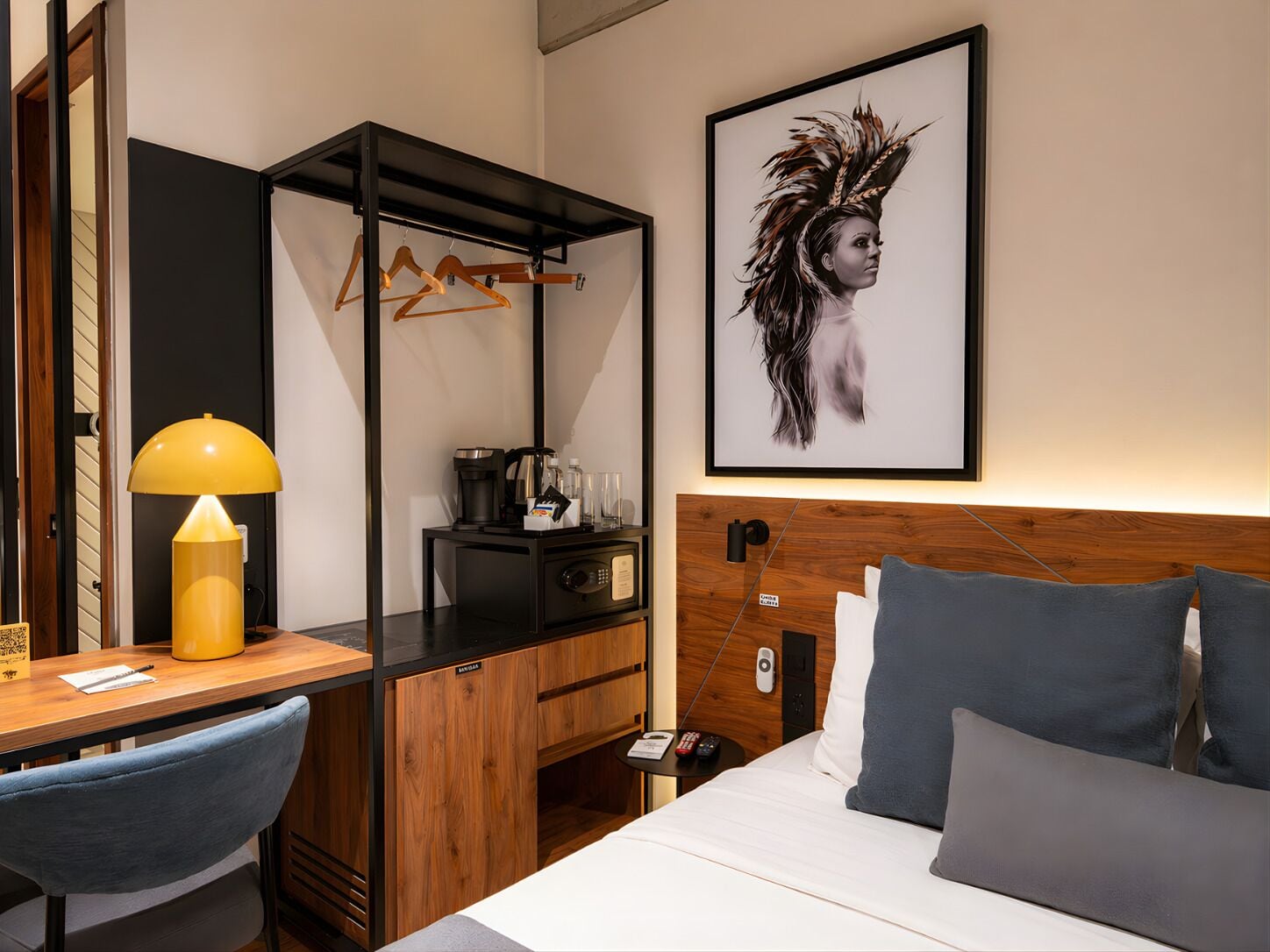 Une chambre d’hôtel moderne équipée d’un lit, d’un bureau avec une lampe, d’une machine à café, d’une armoire et d’une œuvre d’art encadrée accrochée au mur.