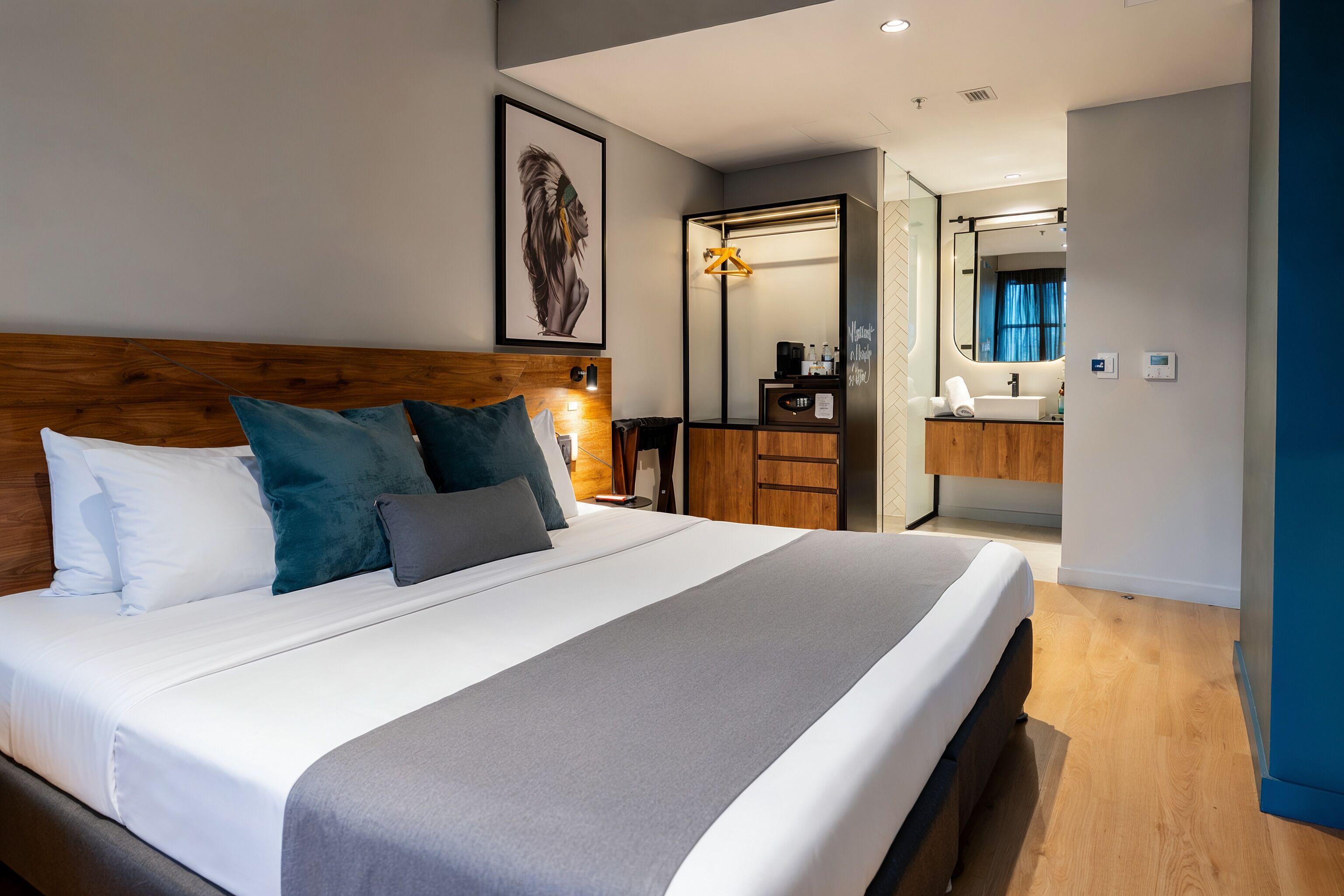 A modern hotel room with a large bed, a wooden headboard, a mirror, a bathroom with a sink, and a framed artwork on the wall.
