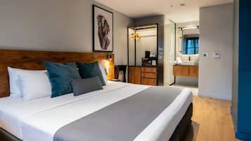 A modern hotel room with a large bed, a wooden headboard, a mirror, a bathroom with a sink, and a framed artwork on the wall.