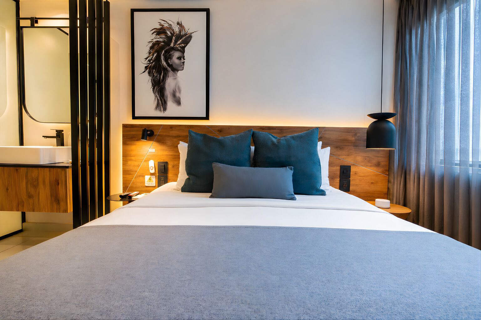 Una habitación de hotel moderna con una cama grande, un cabecero de madera, una mesita de noche y un cuadro en la pared. Se aprecia una ventana con cortinas.