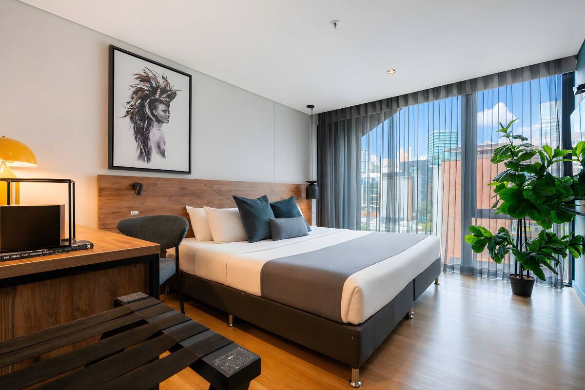 A modern hotel room with a large bed, a desk with a lamp, a chair, a large window with curtains, and a potted plant.