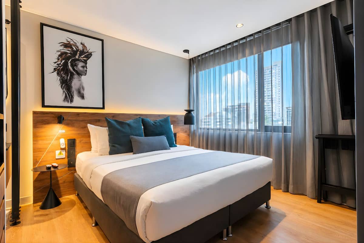 A modern hotel room with a large bed, a headboard with built-in controls, a bedside table with a lamp, and a large window with curtains.