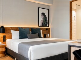 Una habitación de hotel moderna con un cabecero de madera, una cama con ropa de cama blanca y gris, una obra de arte enmarcada sobre la cama, una mesita de noche con una lámpara y un pequeño mueble auxiliar de madera.