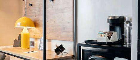 A hotel room with a wooden wall panel, a yellow lamp, a coffee machine, and a small table with a notebook and pen.
