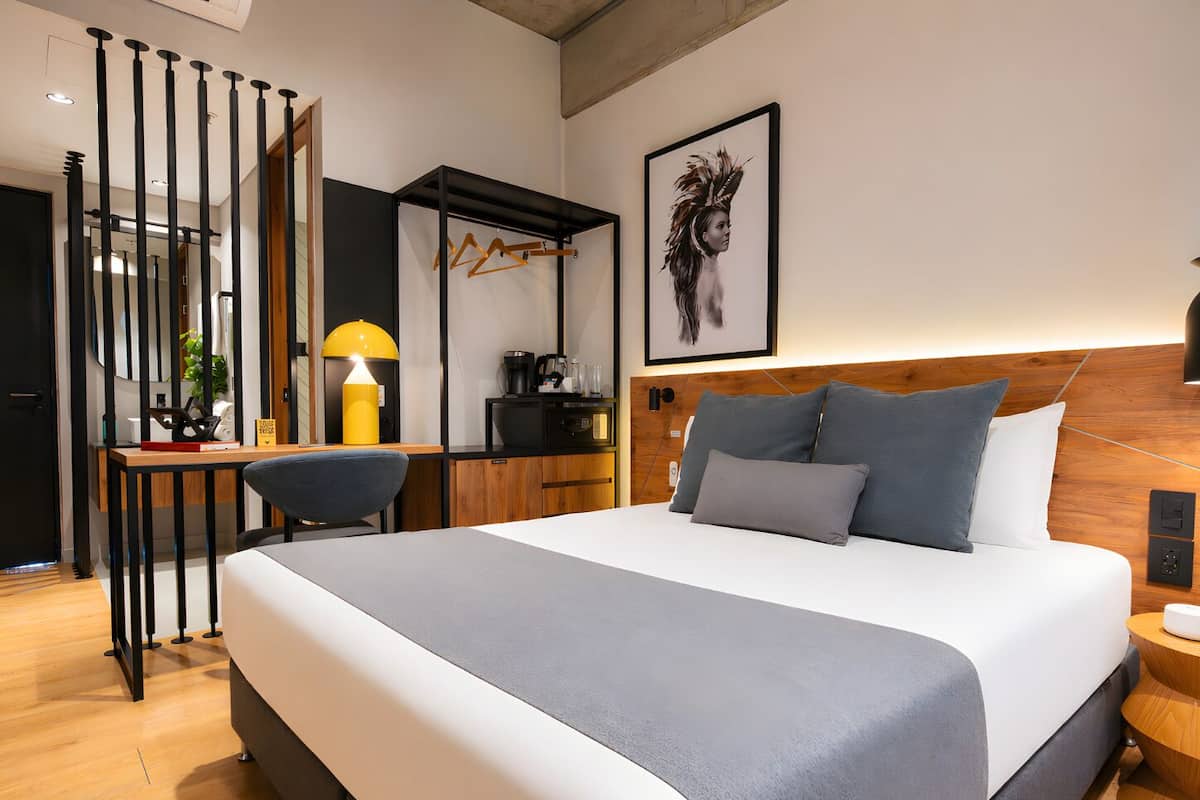 A modern hotel room with a large bed, a desk with a lamp, a wardrobe, and a framed artwork on the wall.