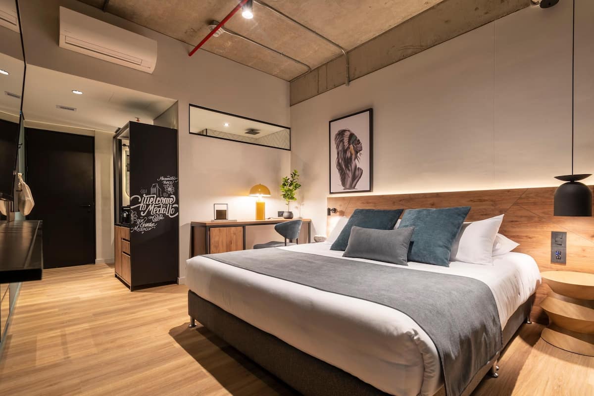 A modern hotel room with a large bed, bedside tables, a desk, a chair, a mirror, and a wall-mounted artwork.