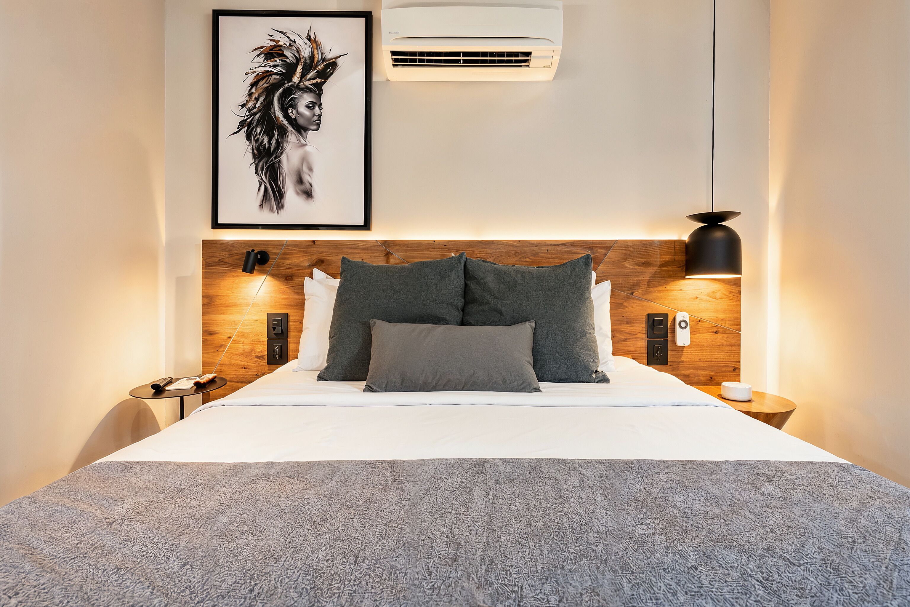 Une chambre d’hôtel moderne avec une tête de lit en bois, une œuvre d’art encadrée, un appareil de climatisation, une lampe de chevet et une table de chevet.