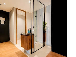A modern hotel room with a wooden floor, a glass partition, and a small wooden desk with toiletries.