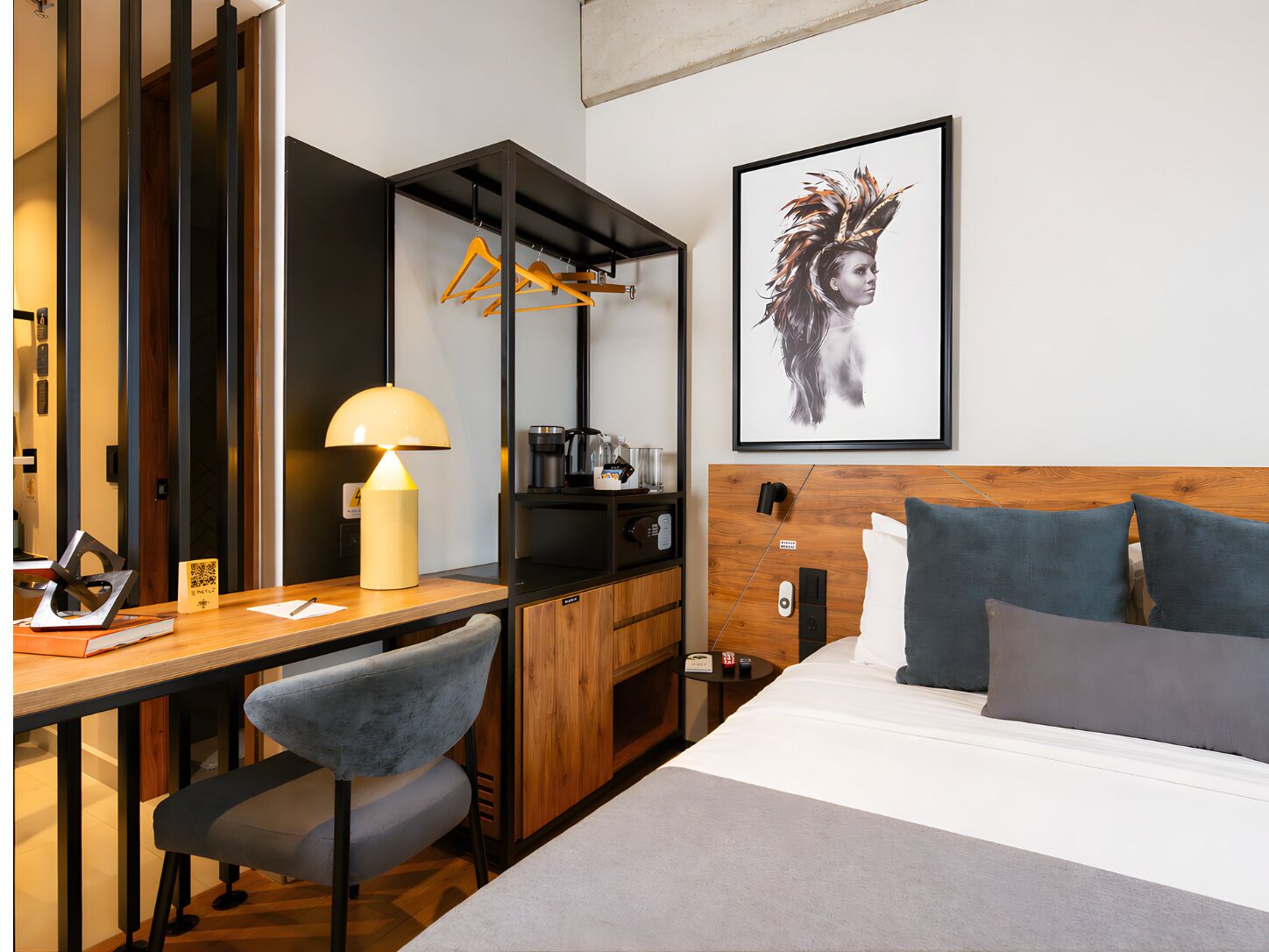 A hotel room with a bed, desk, chair, lamp, and a framed artwork on the wall.