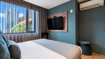 Habitación de hotel con cama, televisor montado, equipo de aire acondicionado y vistas a edificios a través de amplias ventanas.