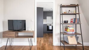 Um espaço de estar moderno com uma TV sobre uma escrivaninha de madeira, uma estante e uma cozinha ao fundo.