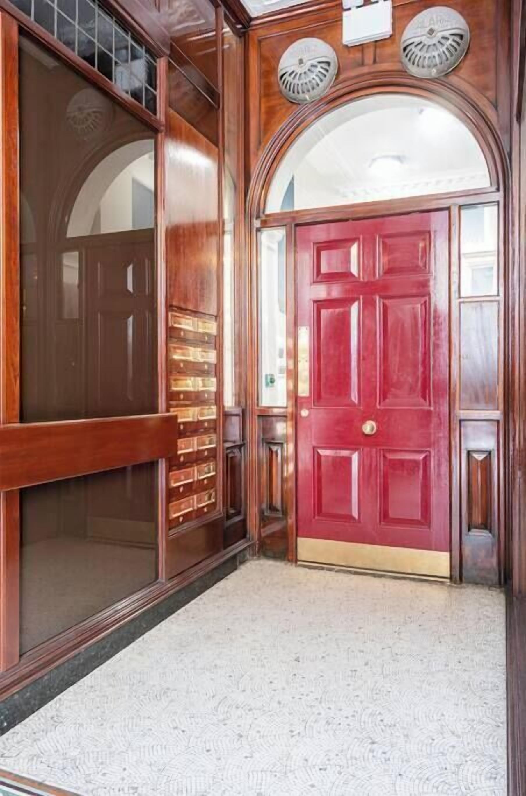 A red door with a brass handle, a wooden wall with a window, and a textured floor.